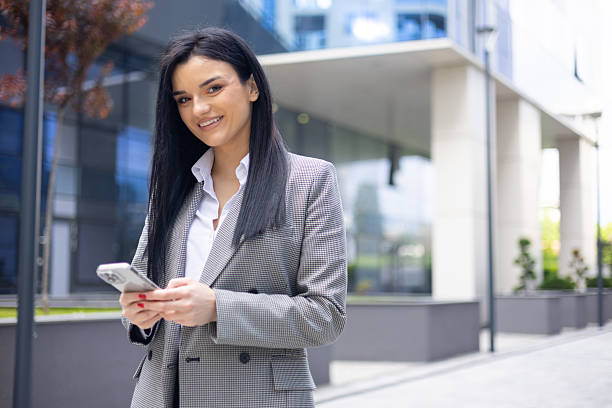 istock Professional businesswoman smiling, checking messages on smartphone while standing near modern office building in urban city 2249695586