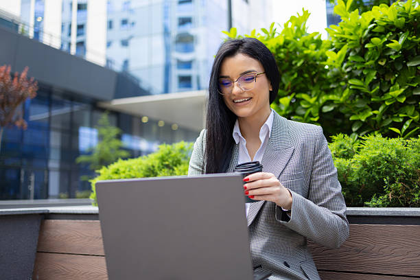 istock Businesswoman enjoying remote work outdoors, using laptop and drinking coffee with city buildings in background 2249698087