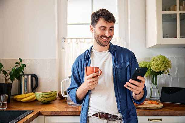 istock Young smiling man using mobile phone while drinking morning coffee at home 2249701116