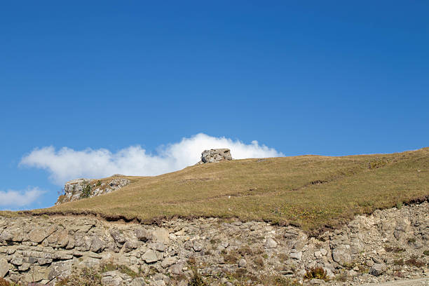 Grassy hill with rocky outcrop and exposed soil under clear blue sky with scattered clouds. Ideal for nature, geology, travel, and commercial use in scenic mountain environments.