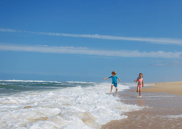 Under a brilliant blue sky, a young boy and girl hold hands run through the foamy waves at the beach happy to spend vacations in sea camp