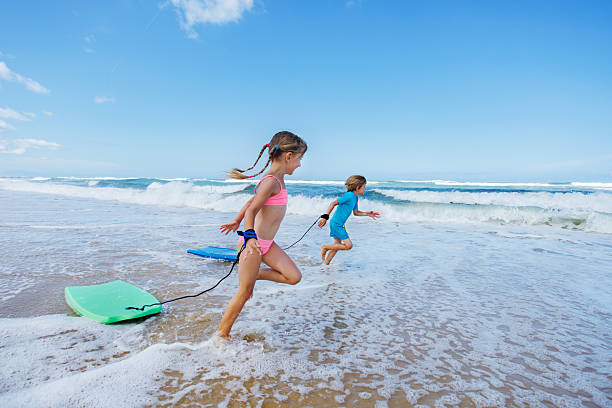 Two children playfully run into the ocean waves with bodyboards on a sunny beach happy to spend vacations in surf school
