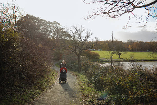 View of the path for walking in the park in autumn. There is a stroller in front of the bench, and a woman with a child is sitting on the bench.