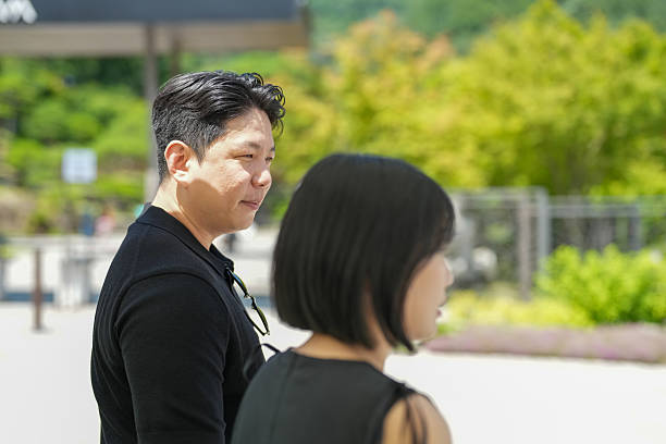 A man and a woman are standing in a park. The man is wearing a black shirt and the woman is wearing a black dress. They are both looking at something in the distance