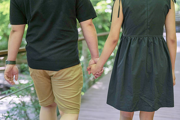 A man and a woman are holding hands while walking on a bridge. The man is wearing a black shirt and the woman is wearing a black dress. Scene is romantic and intimate