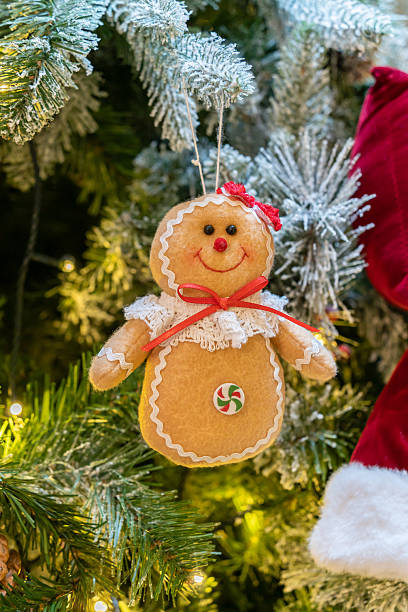 A cheerful gingerbread ornament with icing trim and a red bow hangs among snow dusted Christmas tree branches, illuminated by warm lights.