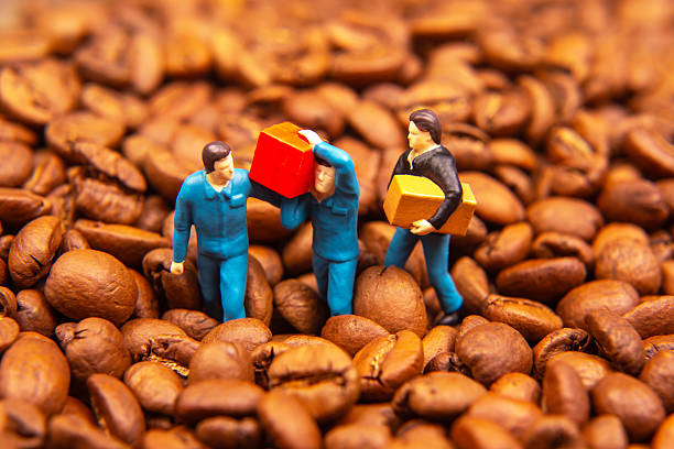 istock A creative close-up of three miniature figures in blue uniforms exchanging a yellow box and a red package amidst a sea of coffee beans, symbolizing trade or commerce. The warm brown tones create a rich, textured background. 2249739640