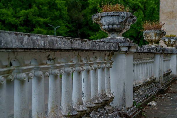 Aged white balustrade with empty stone urn planters lines an abandoned terrace beside lush trees.