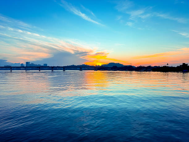 istock A serene sunset over calm waters, with vibrant orange and blue reflections on the surface and a silhouette of a distant bridge and landscape on the horizon. 2249741606