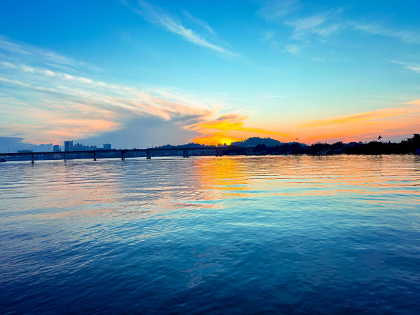 istock A serene sunset over calm waters, with vibrant orange and blue reflections on the surface and a silhouette of a distant bridge and landscape on the horizon. 2249741611
