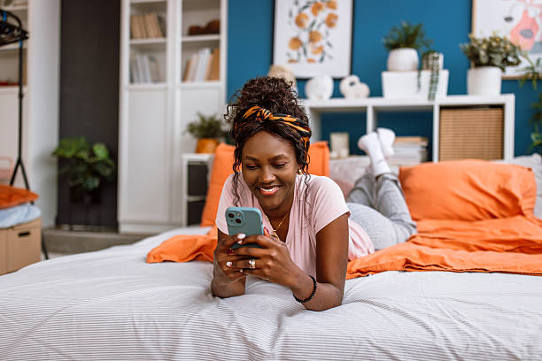 Portrait of a beautiful smiling black woman in pajamas, enjoying the morning at home. She is smiling and using a phone while lying down in her bed.