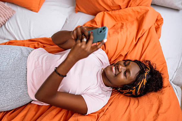 Portrait of a beautiful smiling black woman in pajamas, enjoying the morning at home. She is smiling and using a phone while lying down in her bed.