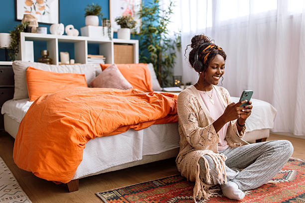 Portrait of a beautiful smiling black woman in pajamas, enjoying the morning at home. She is smiling and using a phone while sitting on the floor in her bedroom.