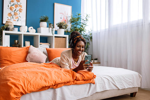 Portrait of a beautiful smiling black woman in pajamas, enjoying the morning at home. She is smiling and using a phone while lying down in her bed.