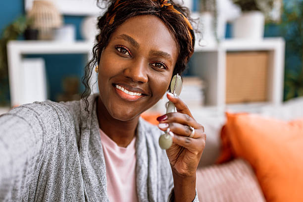 Cheerful mid adult black woman doing routine skin care in bedroom in the morning. Woman using Gua Sha massage face roller.