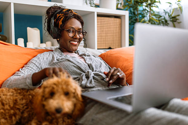 Cheerful mid adult black woman lying on bed, enjoying watching movie on laptop and eating biscuit snack, in company of her cute poodle dog, at home on weekend morning.