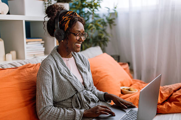 Cheerful mid adult black woman lying on bed, enjoying watching movie on laptop and eating biscuit snack at home on weekend morning.