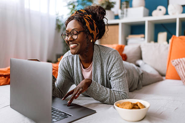 Cheerful mid adult black woman lying on bed, enjoying watching movie on laptop and eating biscuit snack at home on weekend morning.