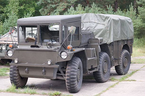 A military transport truck stands parked on a paved surface surrounded by greenery. The vehicle has a canvas cover and rugged tires, typical for off road conditions.