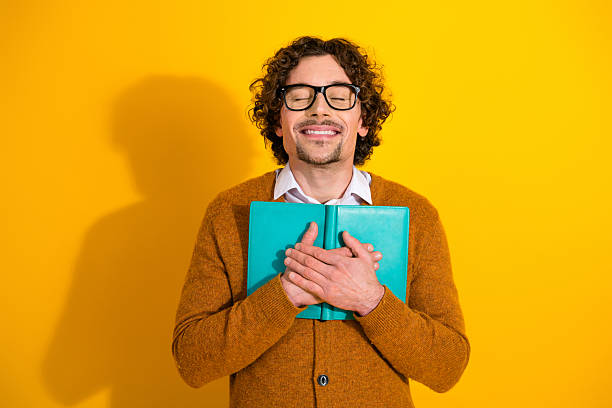 istock Young man with glasses in a yellow studio holding turquoise notebook smiling in a brown cardigan lifestyle portrait 2249757279