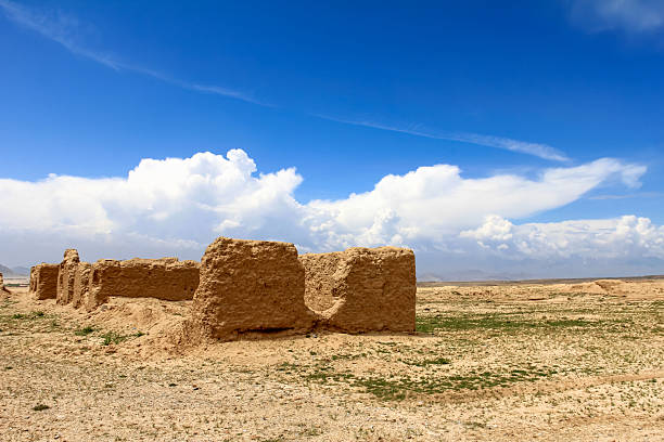 Historic clay ruins stand in an arid desert landscape, framed by clear blue skies and wispy clouds. A depiction of ancient architecture amidst a desolate yet vibrant natural setting, invoking nostalgia and curiosity.