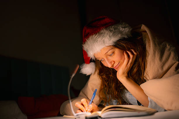 istock Woman wearing a santa hat and comfortable under a blanket, thoughtfully writing her christmas wish list or diary in a notebook on her bed at night 2249760370