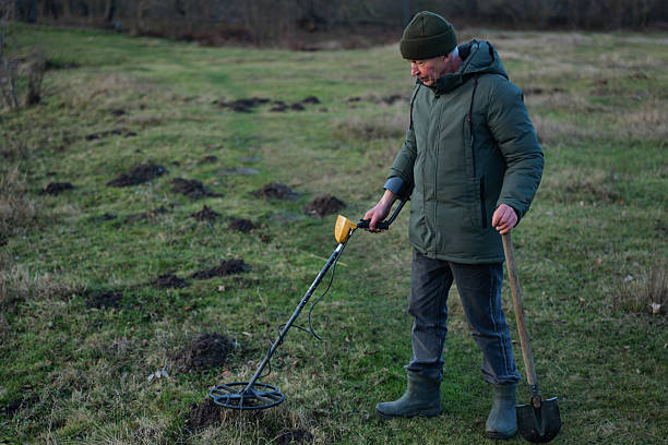 istock A man in a green jacket scans the ground with a metal detector while standing in a grassy field covered in small dirt mounds during early morning light 2249764560