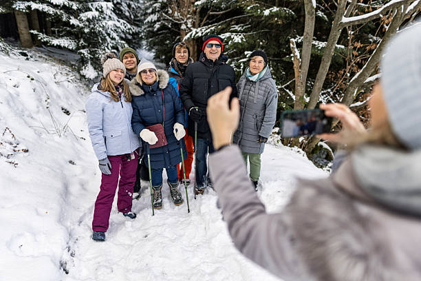 Group of people stand close together in a snowy forest, smiling for a photo taken by a seventh companion with a smartphone. Dressed in winter jackets, hats, gloves, and boots, some hold walking poles, they enjoy a shared hike through the cold. Snow blankets the ground and trees, creating a serene backdrop for this moment of pause and connection.Shot with Canon R5.