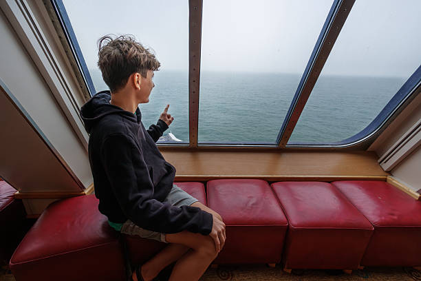 A young person traveler in a hoodie sits on red benches, gazing at the ocean through a ship's window, while gesturing towards the horizon