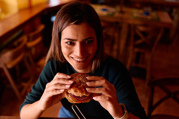Young adult woman expressing joy and anticipation, holding a delicious burger with both hands while sitting at a wooden table in a restaurant setting, ready to enjoy her meal