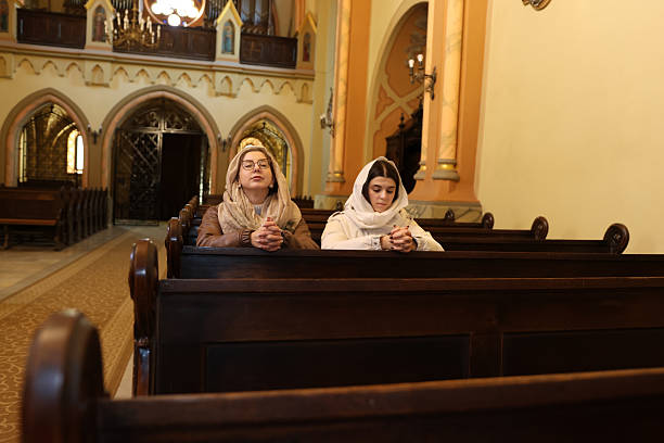 Two women sit on a bench in a church, focused on prayer.