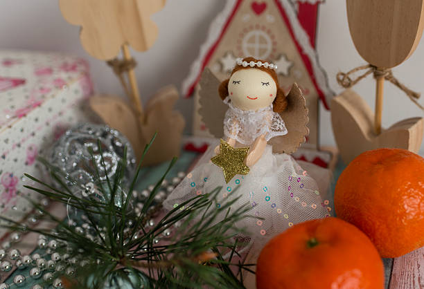 A cute Christmas angel doll with a gold star sits among ornaments, pine branches, beads and mandarins. A warm and charming holiday scene ideal for winter greeting cards, promotions, and family-themed content.