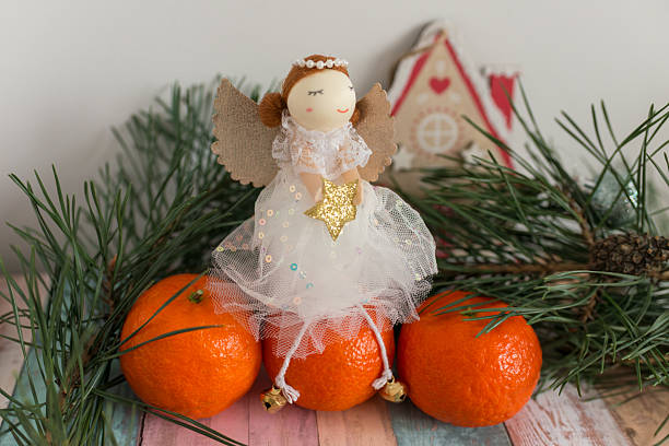 A decorative Christmas angel sits above bright mandarins and pine branches, creating a festive seasonal still life. Ideal for holiday greetings, winter marketing, gift designs, and New Year visuals.