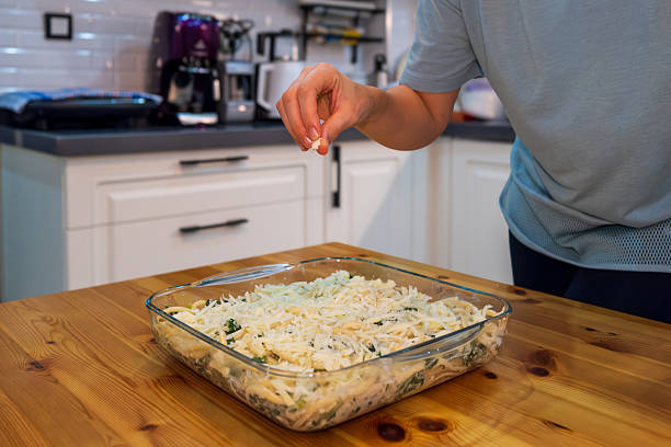 A person sprinkles shredded cheese over a homemade casserole in a glass baking dish, preparing it for the oven. The scene takes place in a bright home kitchen with modern cabinetry. The image captures the process of meal preparation, highlighting themes of home cooking, comfort food, and culinary routines.