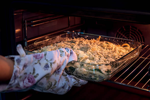 A person wearing floral oven mitts places an uncooked casserole into a preheated oven. The glass baking dish is topped with shredded cheese, ready to be baked. This image conveys themes of home cooking, preparation, kitchen routines, and homemade meals.