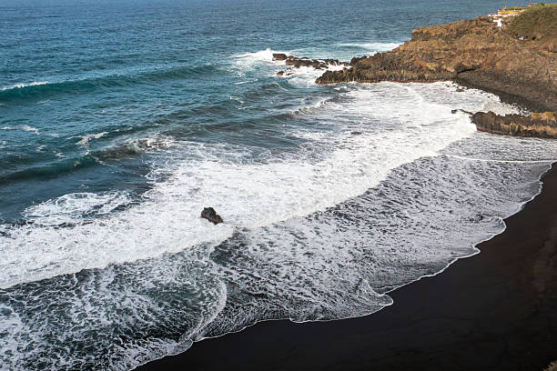 A dramatic, wide-angle view of Playa El Bollullo, a secluded black sand beach on the wild, northern coast of Tenerife, Canary Islands, Spain. The scene captures the raw, untouched natural beauty with powerful Atlantic waves crashing against the shoreline, contrasting with the dark volcanic sand and the sheer cliffs of the La Orotava Valley. This spot is renowned for its untamed surf and tranquil, natural atmosphere.