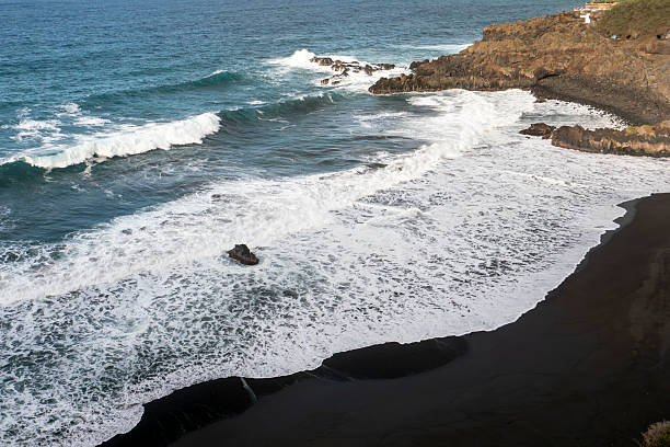 A dramatic, wide-angle view of Playa El Bollullo, a secluded black sand beach on the wild, northern coast of Tenerife, Canary Islands, Spain. The scene captures the raw, untouched natural beauty with powerful Atlantic waves crashing against the shoreline, contrasting with the dark volcanic sand and the sheer cliffs of the La Orotava Valley. This spot is renowned for its untamed surf and tranquil, natural atmosphere.