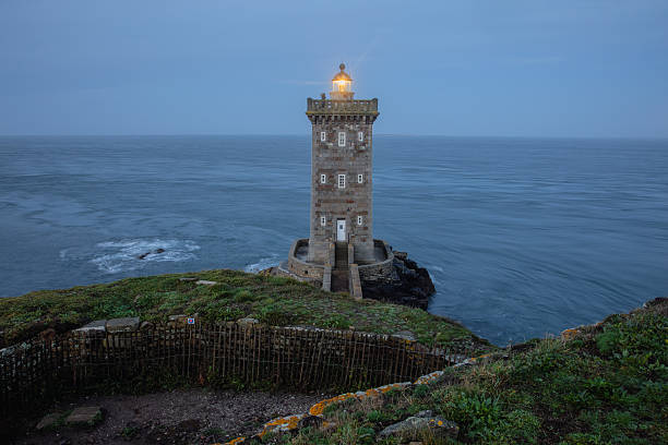 A serene blue-hour view of the historic Kermorvan Lighthouse standing on a rugged headland in Le Conquet, Brittany, France. The warm glow of the lantern contrasts with the cool tones of the Atlantic Ocean, as gentle long-exposure waves soften the surrounding seascape. Ancient stone walls, coastal vegetation and dramatic cliffs frame the scene, highlighting the lighthouse’s vital role in maritime navigation and its iconic presence on the westernmost edge of mainland France