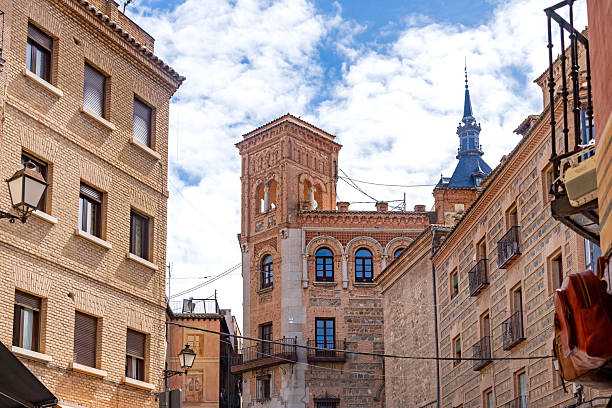 A patterned brick corner building in Toledo with iron balconies and bay windows beside a historic bell tower with arched openings under a blue, cloudy sky