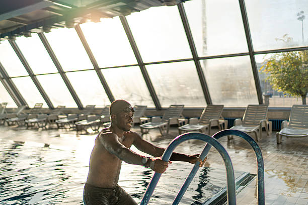 Young black man smiling as he climbs out of a modern indoor pool, enjoying aquatic exercise and leisure in sunlit, glass-walled surroundings that evoke fitness and relaxation