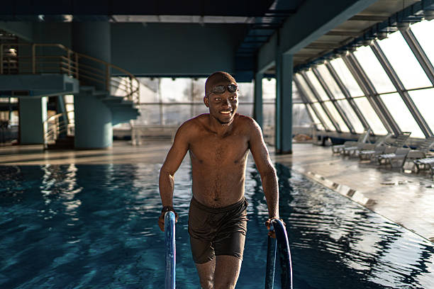 Smiling african american man wearing swimming goggles and trunks, looking away while coming out of a vibrant indoor pool, enjoying recreational sport and exercise