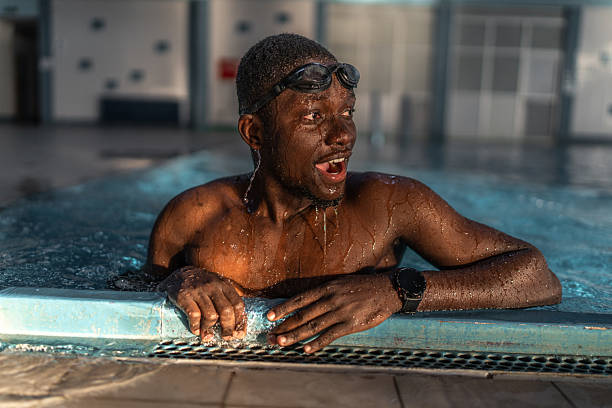Young african man in swim goggles smiling while leaning on the pool edge, wet hair dripping, enjoying a refreshing indoor swim and carefree summer fitness moment