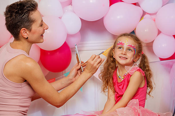 In front of pink balloons, a woman is face painting a child with bright hues colors, kids birthday party celebration