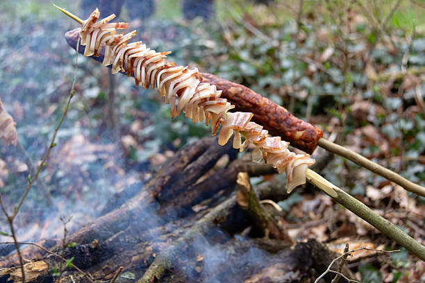 Tasty sausages and bacon skewered on sticks are roasted over a burning campfire in a rustic forest setting, creating a warm and traditional outdoor cooking atmosphere surrounded by nature.