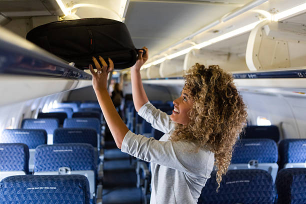 istock Woman stowing carry-on bag in overhead compartment on airplane with smiling expression. 2249834479
