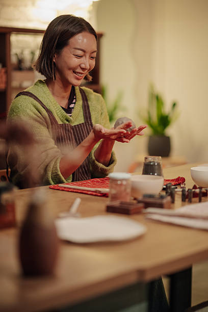 Asian woman smiling, holding and shaping a small piece of clay with her hands at a creative pottery workshop