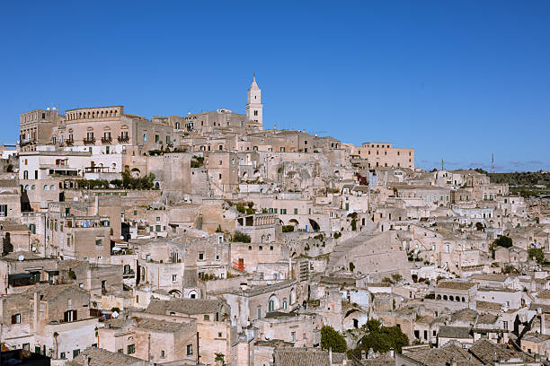 panoramic view of matera's sassi, an unesco world heritage site, featuring historic cave dwellings and the iconic bell tower under a bright blue sky. a testament to ancient architecture and history