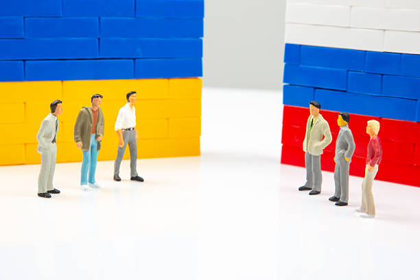 Conceptual photo featuring small human figures separated by blocks walls painted with Ukraine and Russia flag colors, on a plain white background.