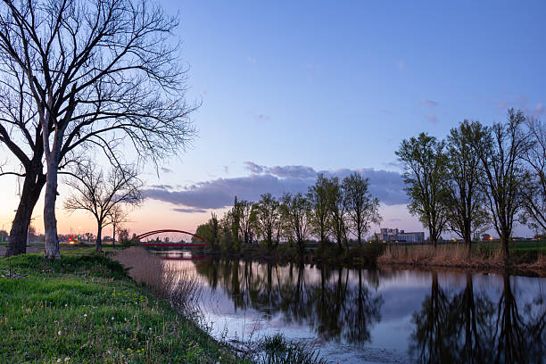 Serene riverside view with a calm water surface showing clear reflections, lined by trees under a peaceful evening sky, featuring a distinctive arched bridge in the background.