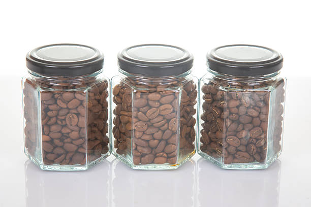 three glass jars with black lids filled with roasted coffee beans arranged in a row on a reflective surface on a white background